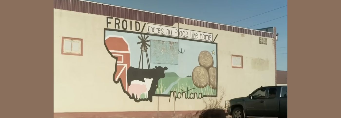 A mural on a building in Froid, Montana reads "there's no place like home" with farm imagery and the state outline.